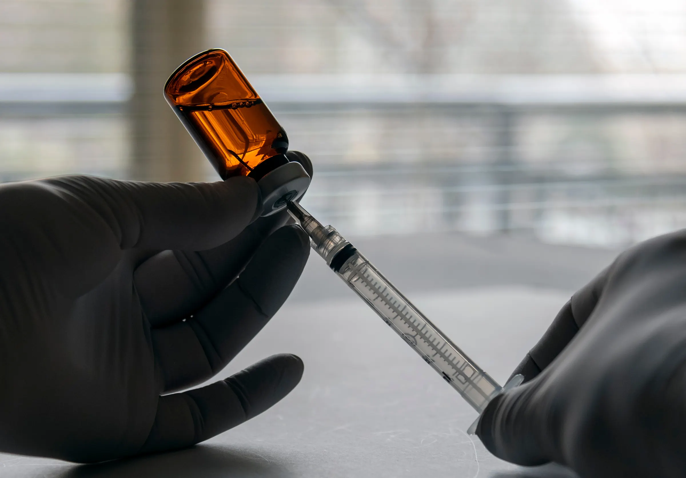 Gloved hands drawing liquid from an amber vial in a clean preparation area